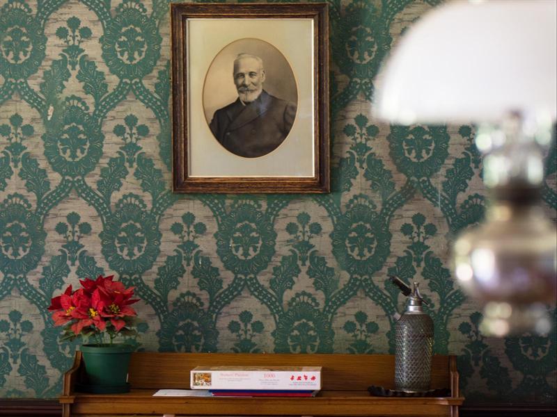 A framed portrait on green damask wallpaper above a side table with flowers and an oil lamp
