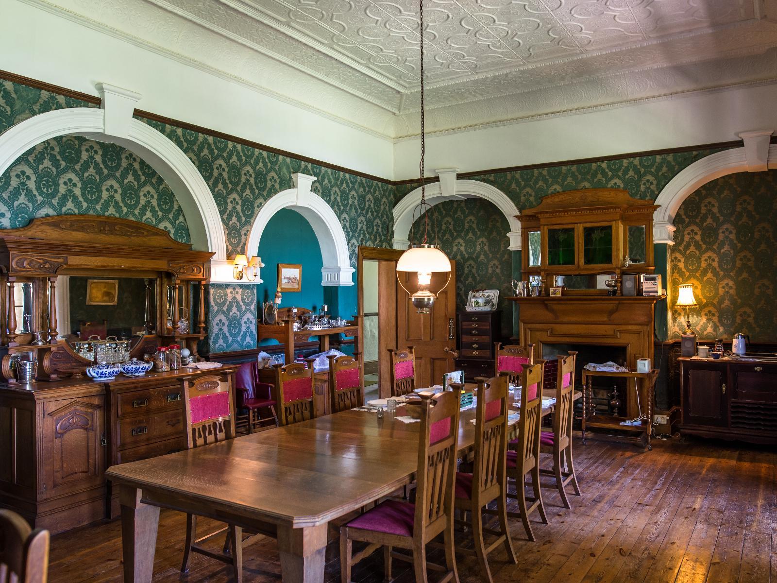 The dining room: a long wooden table under a pressed ceiling, framed by arched alcoves, green damask wallpaper and a fireplace